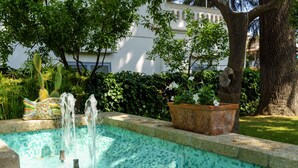 Una piscina con fuente, rodeada de vegetación y un edificio al fondo.