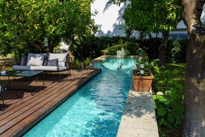 A swimming pool with a wooden deck, a lounge area with a sofa and table, and a water feature.