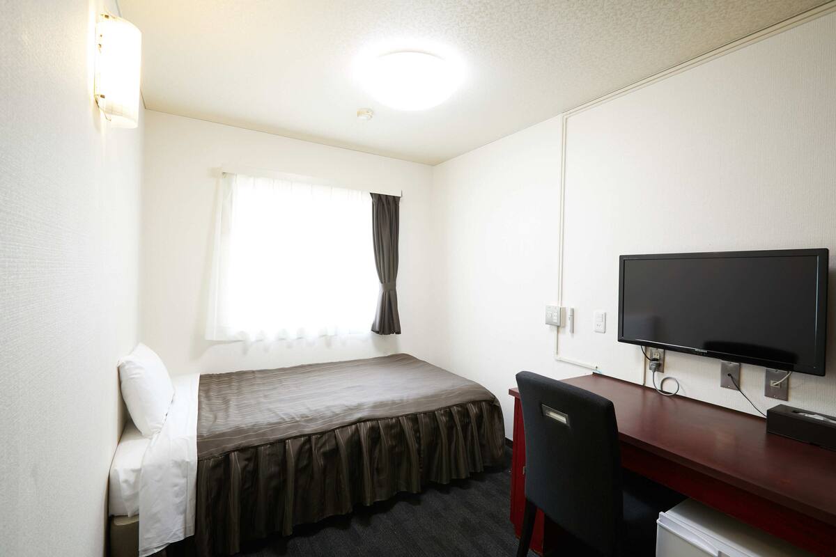A small, clean room with a bed, a desk, a chair, a TV, and a window with curtains.