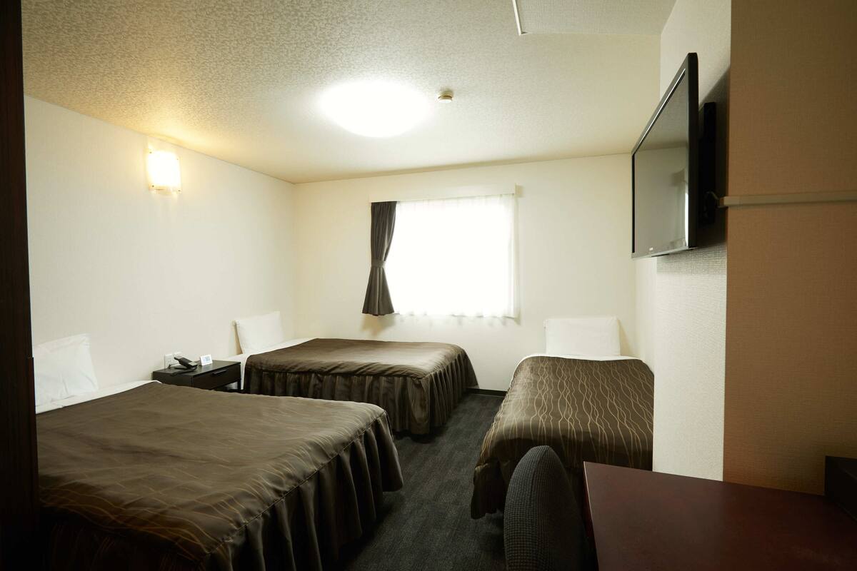 A hotel room with two beds, a desk, a chair, a flat-screen TV, and a window with curtains.
