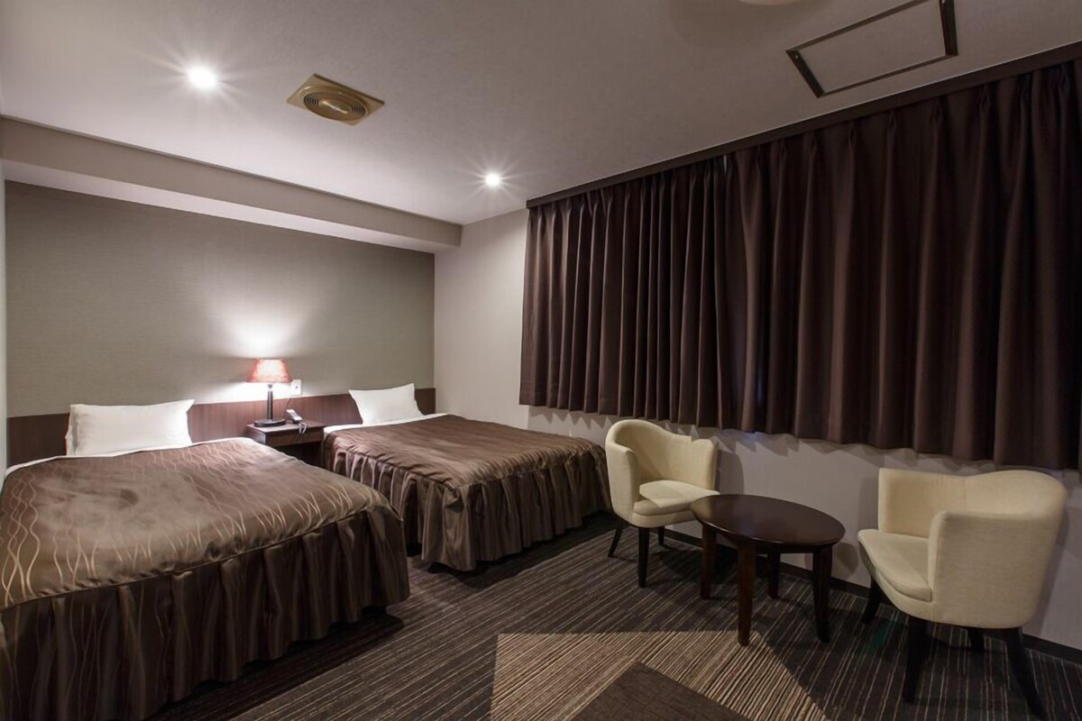 A hotel room with two beds, a desk, a chair, and a window with curtains.