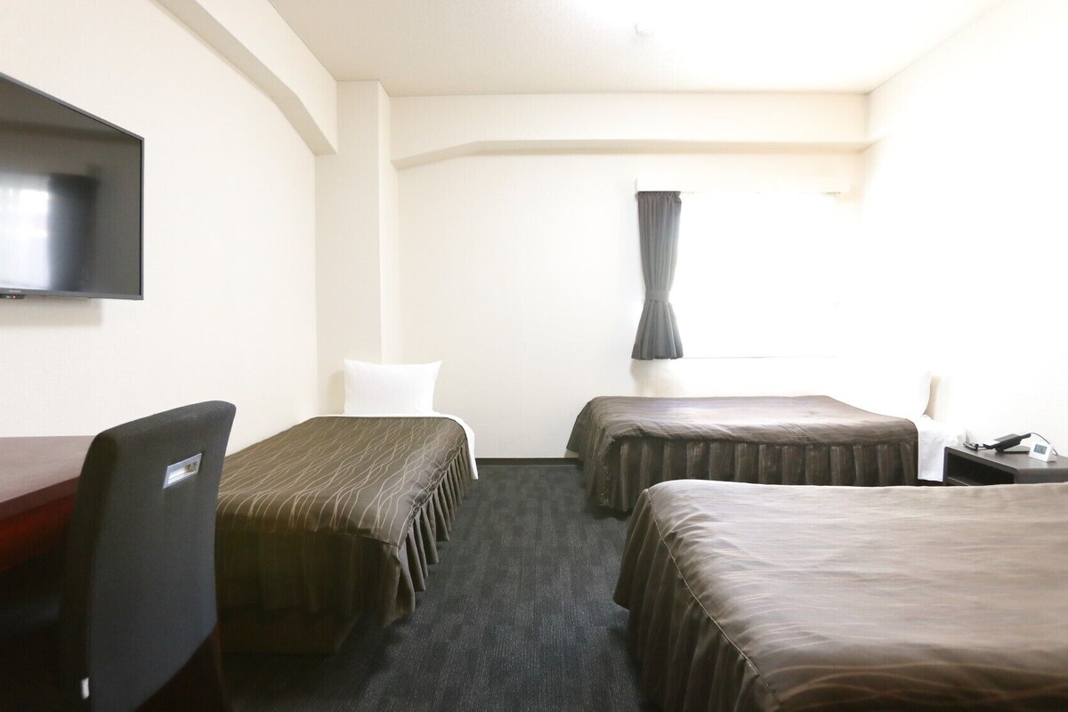 A hotel room with a desk, two beds, a TV, and a window with curtains.