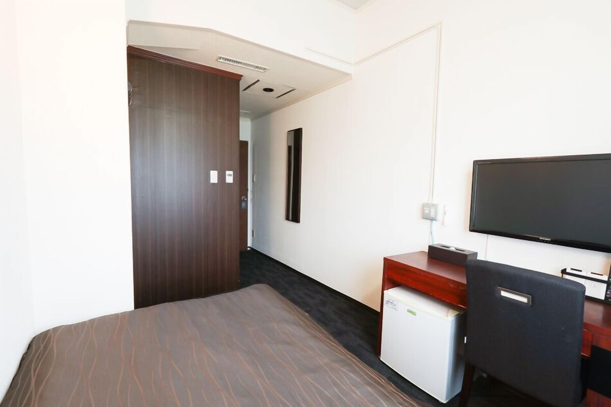 A hotel room with a bed, a desk with a television, a small refrigerator, and a door leading to another room.