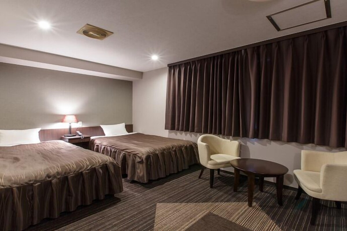 A hotel room with two beds, a desk, a chair, and a large window with curtains.