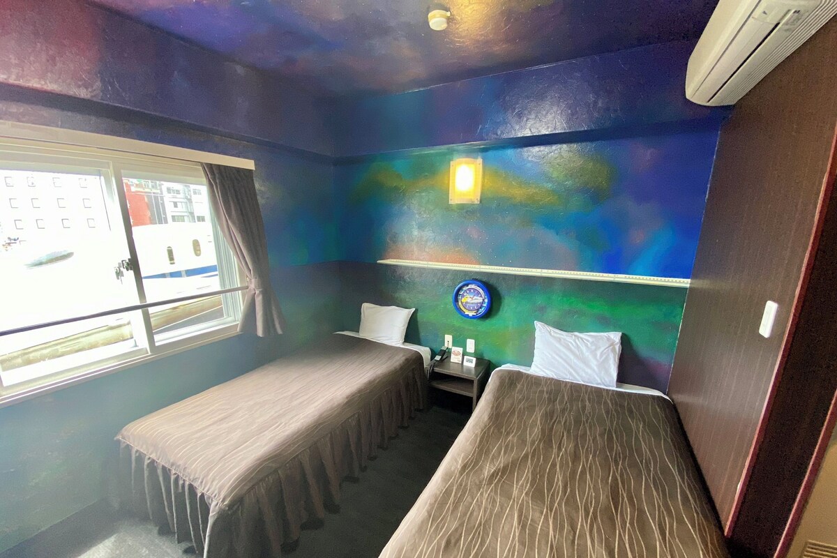A room with two single beds, a window with curtains, a wall clock, and a small table with a lamp.