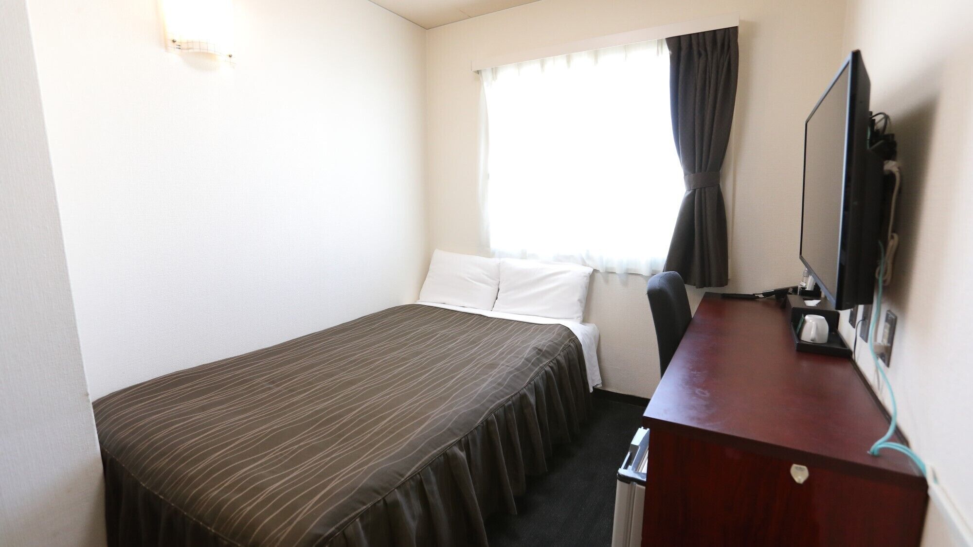 Quarto de hotel com uma cama de solteiro, escrivaninha com cadeira, televisão e janela com cortinas.