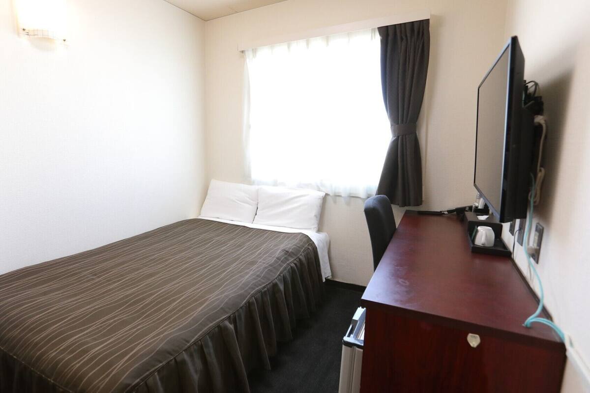 A hotel room with a single bed, a desk with a chair, a television, and a window with curtains.