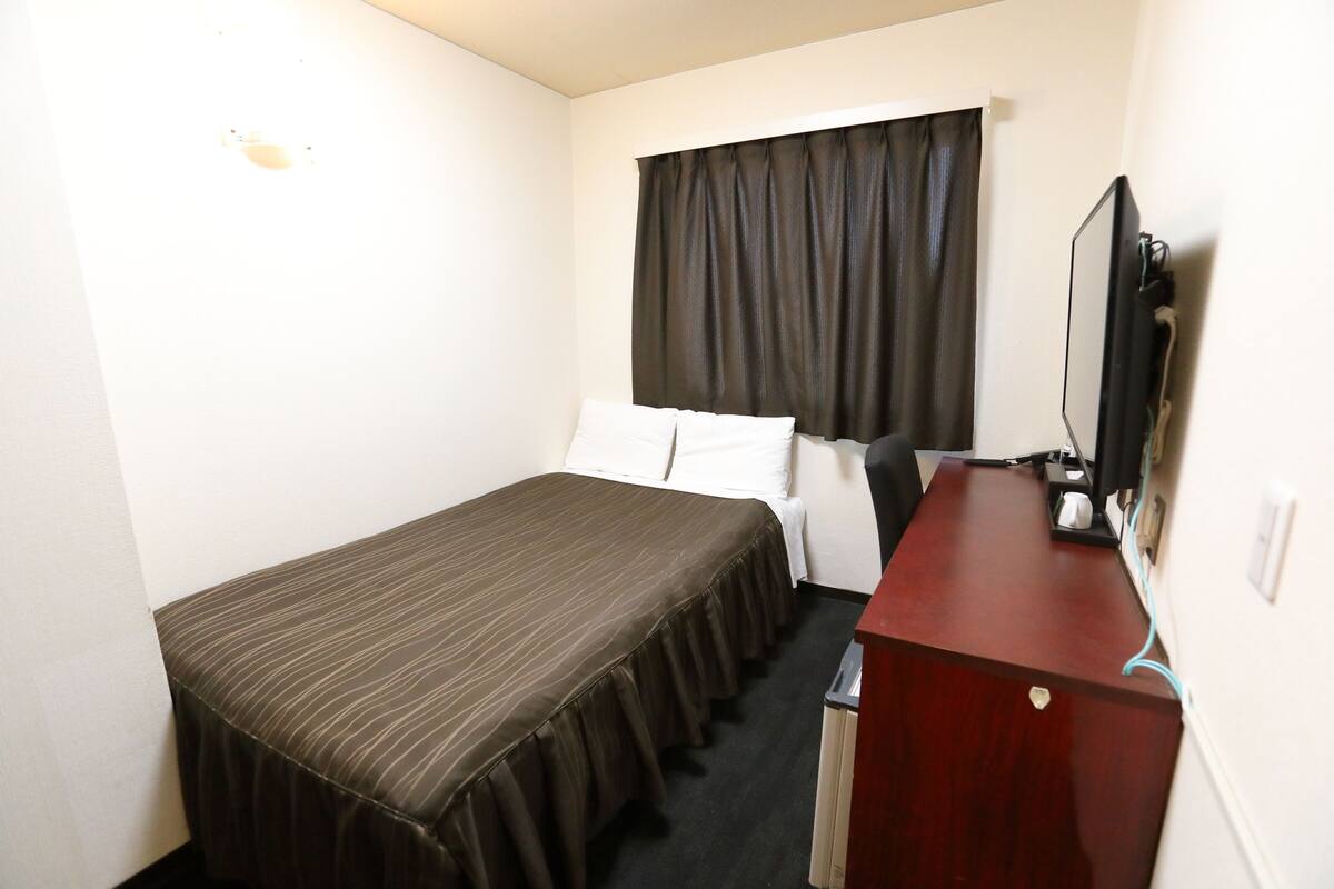 A small hotel room with a single bed, a desk with a television, and a window with curtains.