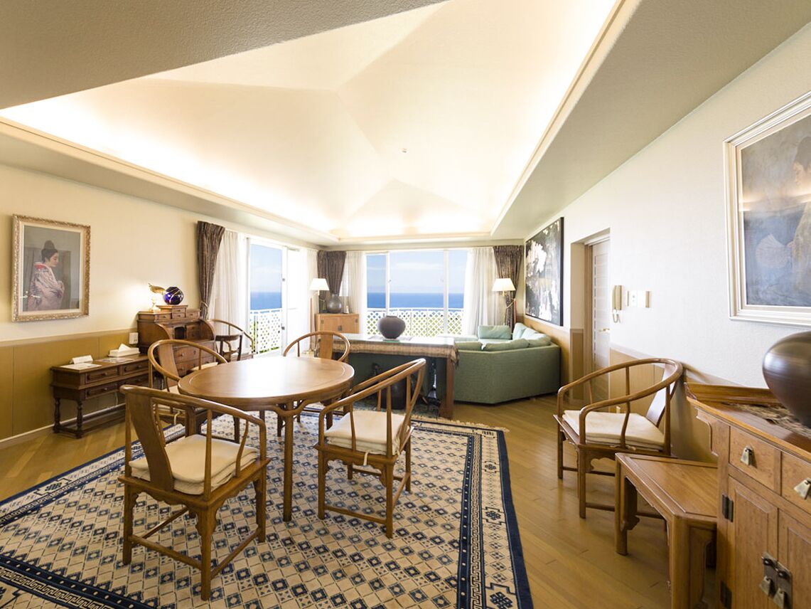 A spacious room with a round dining table, wooden chairs, a green sofa, and a view of the ocean through large windows.