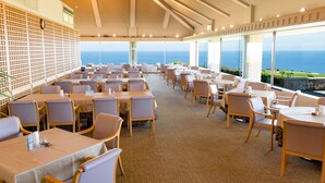 A spacious dining area with tables and chairs arranged for guests, large windows offering ocean views, and a ceiling with a geometric pattern. - Southern links resort hotel (Yaese)