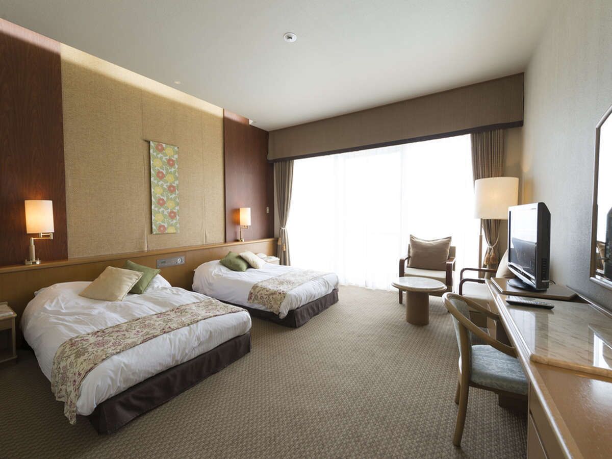 Ein Hotelzimmer mit zwei Betten, einem Schreibtisch mit Stuhl, einem Fernseher und einem großen Fenster mit Vorhängen.