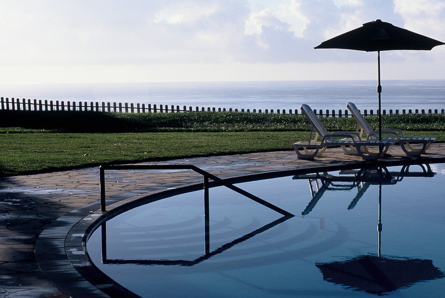 Piscina com espreguiçadeiras e um guarda-sol, cercada por uma vedação e vegetação.