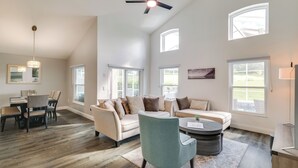 Ein geräumiges Wohnzimmer mit hoher Decke, Deckenventilator und großen Fenstern für natürliches Licht. Es gibt einen Essbereich mit Tisch und Stühlen sowie eine Sitzecke mit Sofa und Sesseln.
