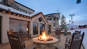 An outdoor seating area with a stone fire pit surrounded by wooden chairs, a building with a snow-covered roof, and a ski lift in the background. - CX113 | Onsite Check-In | Direct Access to Ski Area | Winter Shuttle | Hot Tub (Steamboat Springs)