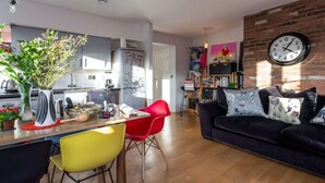 Uma sala de estar moderna com um sofá escuro, cadeiras vermelhas e uma área de cozinha com geladeira e relógio na parede.