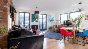 Uma sala de estar moderna com um sofá preto, uma mesa de jantar com cadeiras coloridas e janelas amplas.
