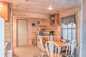 A compact kitchen with wooden walls, a microwave, stove, and white chairs around a table.