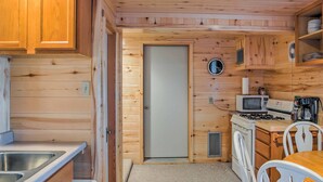 Cocina pequeña con paredes de madera, microondas, estufa, lavabo y sillas blancas.