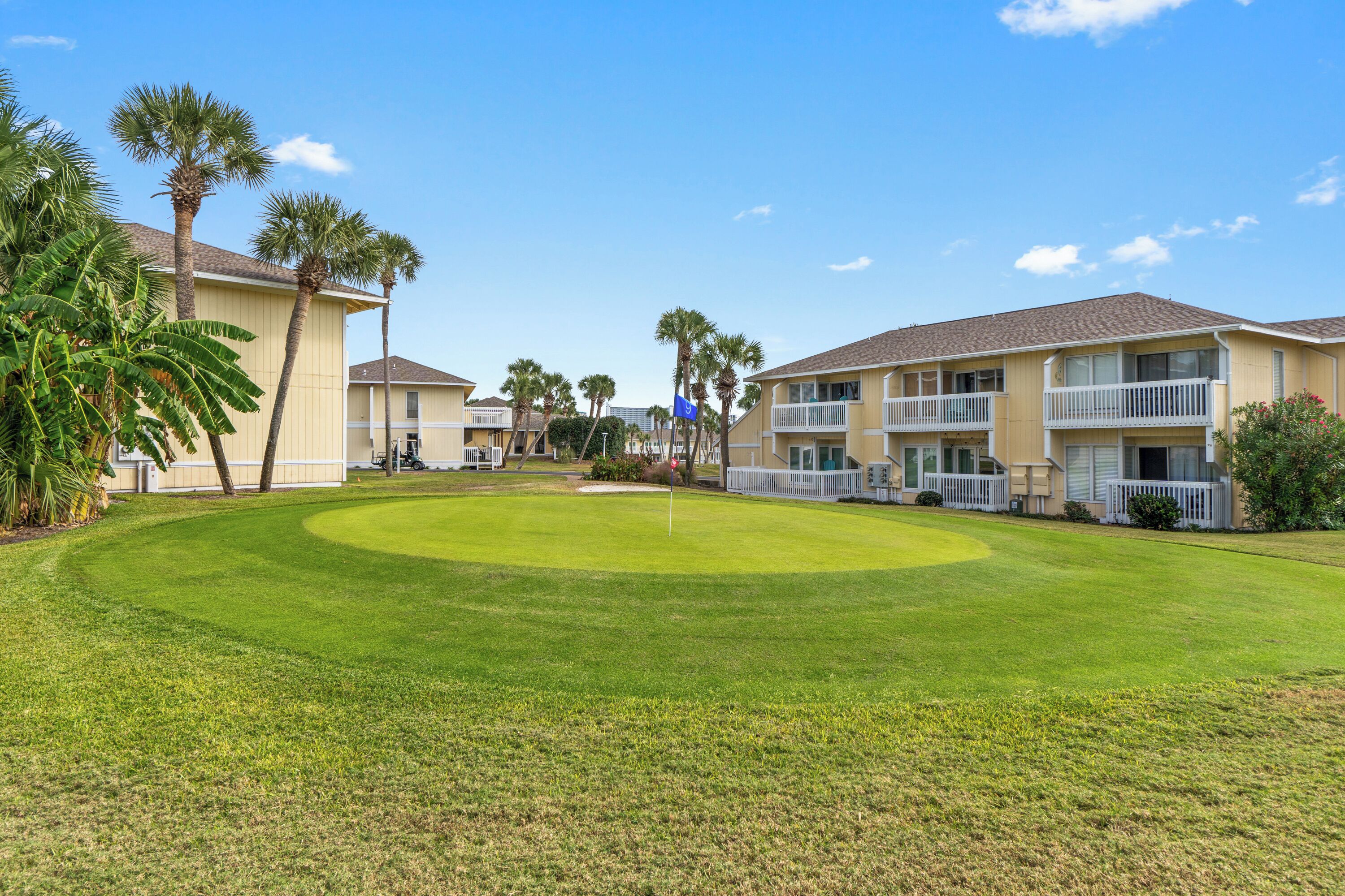 A golf course with a flagstick, surrounded by palm trees and a building with balconies.