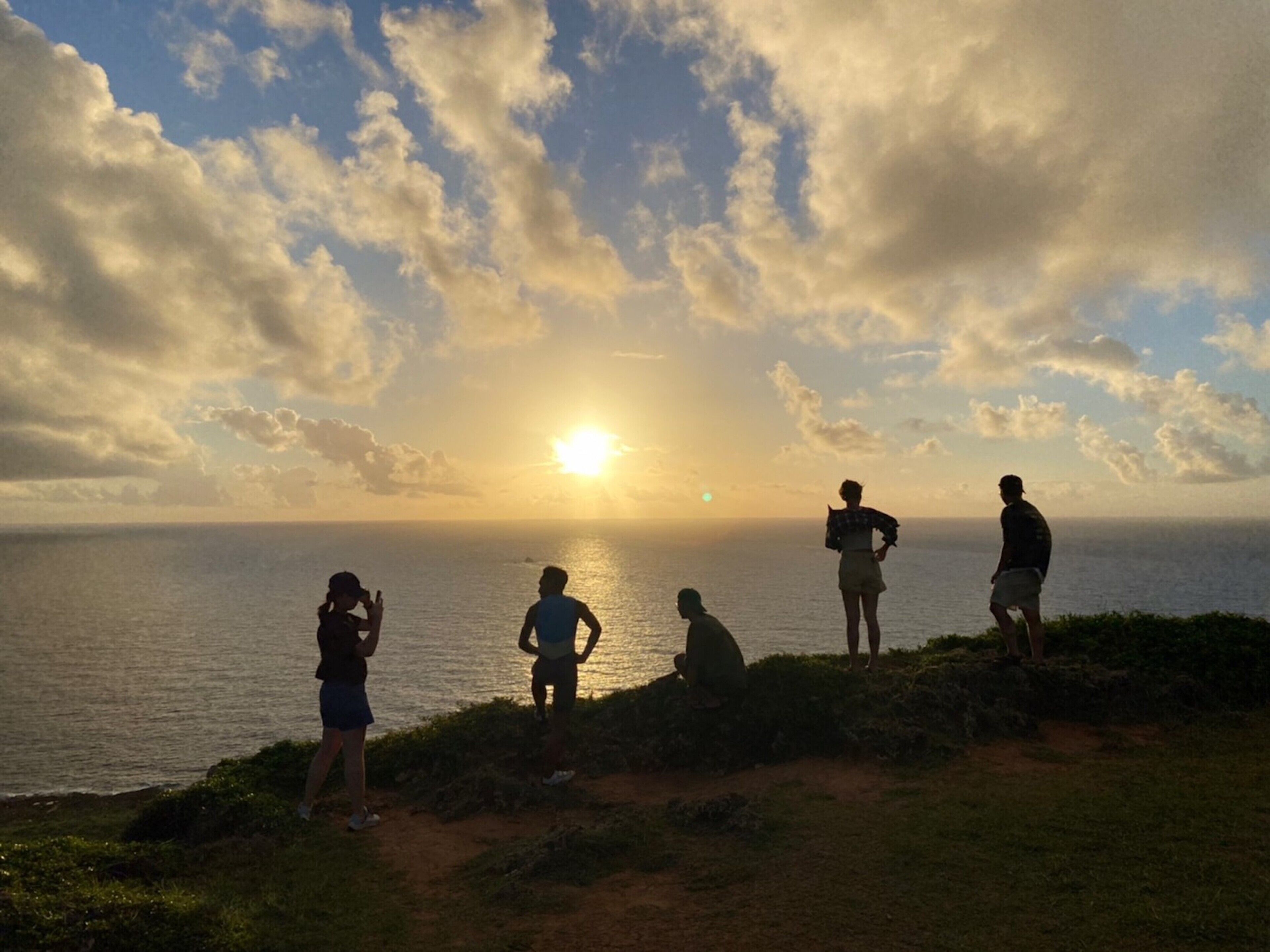 A sunset view with people standing on a grassy area overlooking the sea.