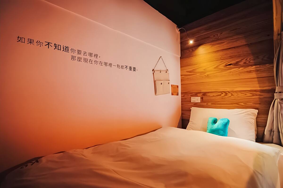 Habitación de hotel con pared de madera, cama con ropa blanca y almohada azul, y una lámpara de pared.