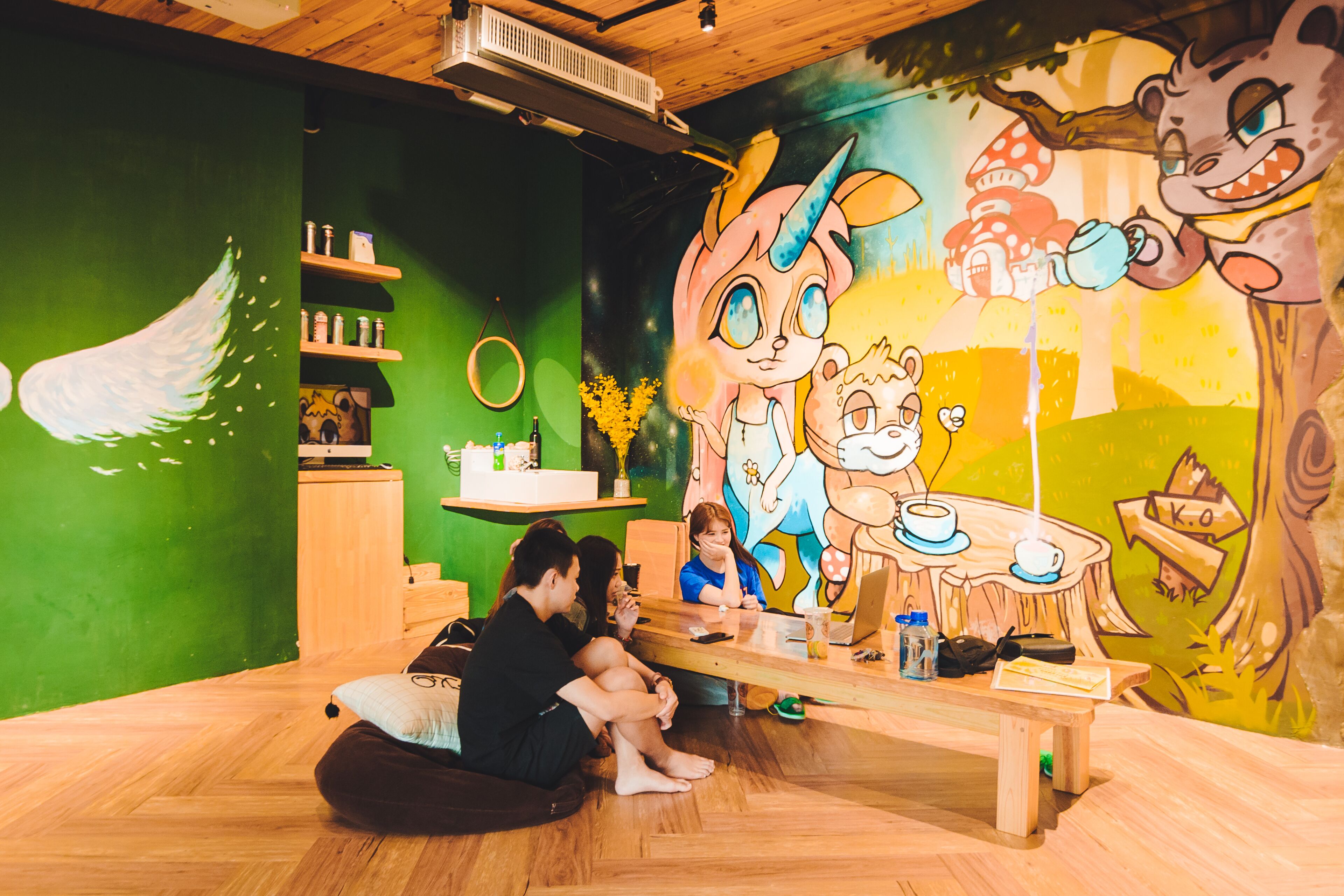 Una habitación con piso de madera, pared verde y un mural de personajes de caricatura. Hay una mesa con una computadora portátil, una botella y una taza.