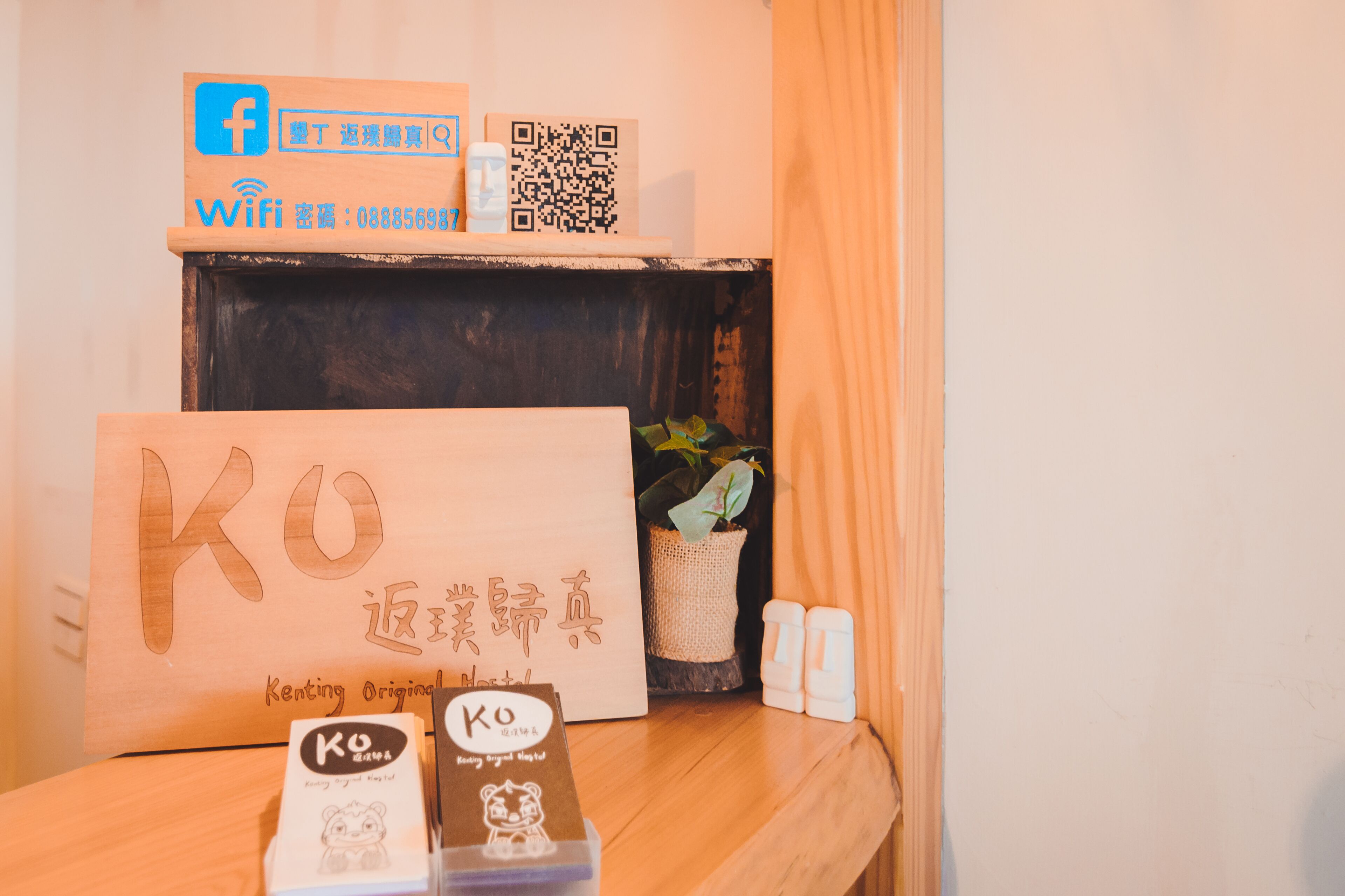 A wooden shelf with a sign reading "KO" and a QR code, a small potted plant, and a couple of white bottles.