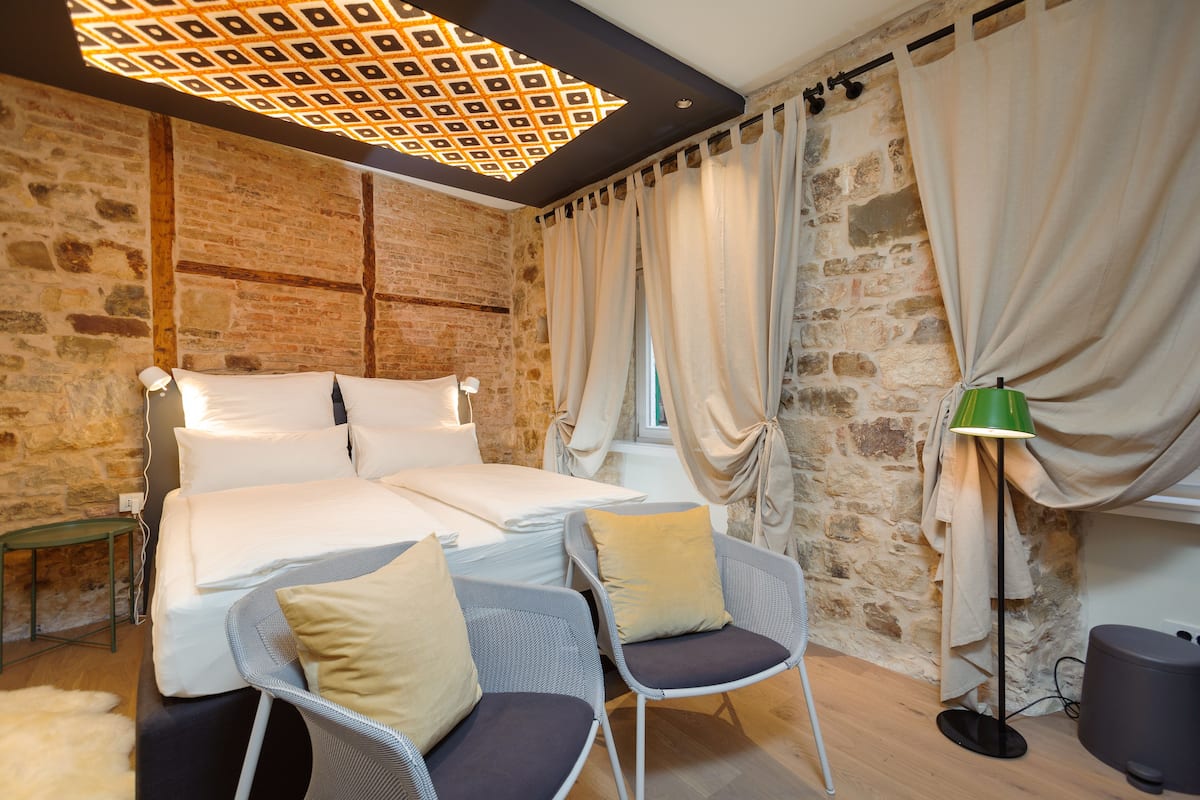A bedroom with a large bed, two chairs, a lamp, and a stone wall.