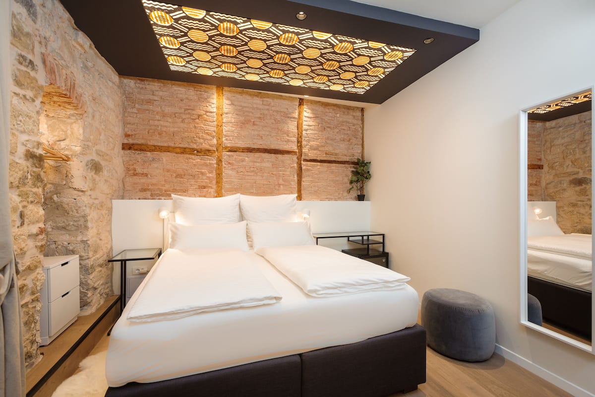 A modern bedroom with a brick wall, a large bed, bedside tables, a mirror, and a unique ceiling light fixture.