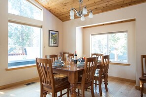 A dining area with a wooden table set for six, surrounded by six wooden chairs. There are two large windows providing natural light and a view of trees outside.