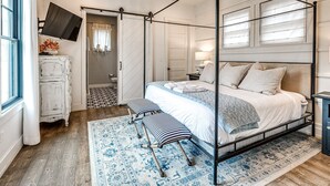 Una cama con dosel con una banca a juego, una araña y una alfombra estampada en un dormitorio con piso de madera y un aparador blanco.