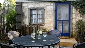 Un edificio de piedra con una puerta azul y un ventanal con cortinas. Hay una mesa redonda con sillas y una botella de vino.