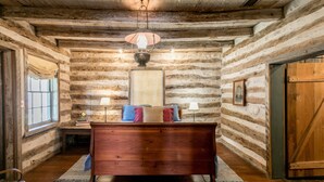 Interior de una cabaña de troncos con una cama de madera, dos lámparas y un cuadro en la pared.