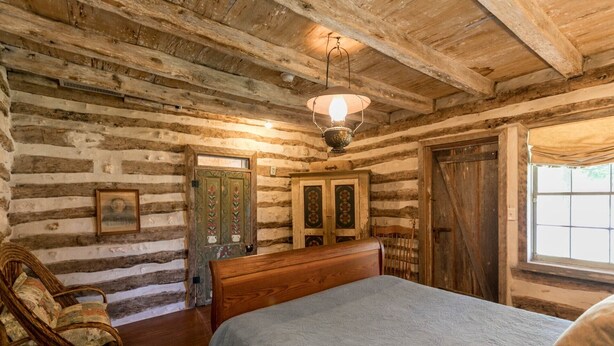 Un dormitorio rústico de cabaña de madera con una cama, una silla, un aparador y una ventana.