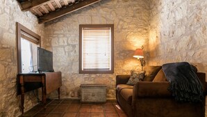 Una habitación acogedora con pared de piedra, techo de madera, un televisor sobre un mueble de madera, un sofá con cojines, un pequeño cofre de madera y una ventana con persianas.
