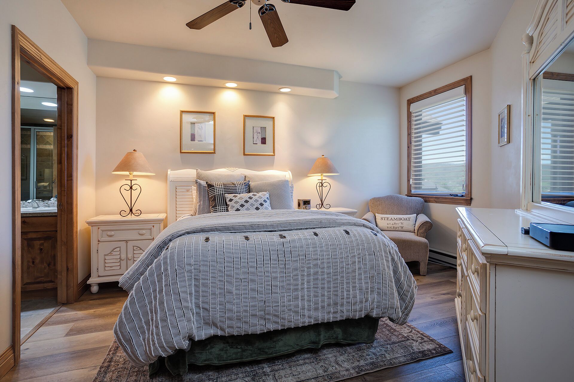 A bedroom with a bed, two bedside tables, a chair, a window with blinds, and a ceiling fan.