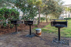 Outdoor barbecue area with three grills, surrounded by lush greenery and a paved patio.