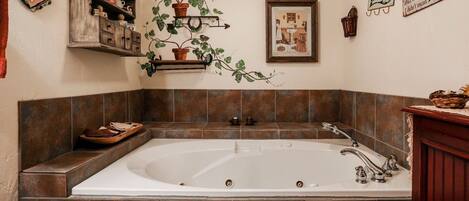 Un baño con una bañera grande, paredes con azulejos, un estante de madera con adornos y un cuadro en la pared.