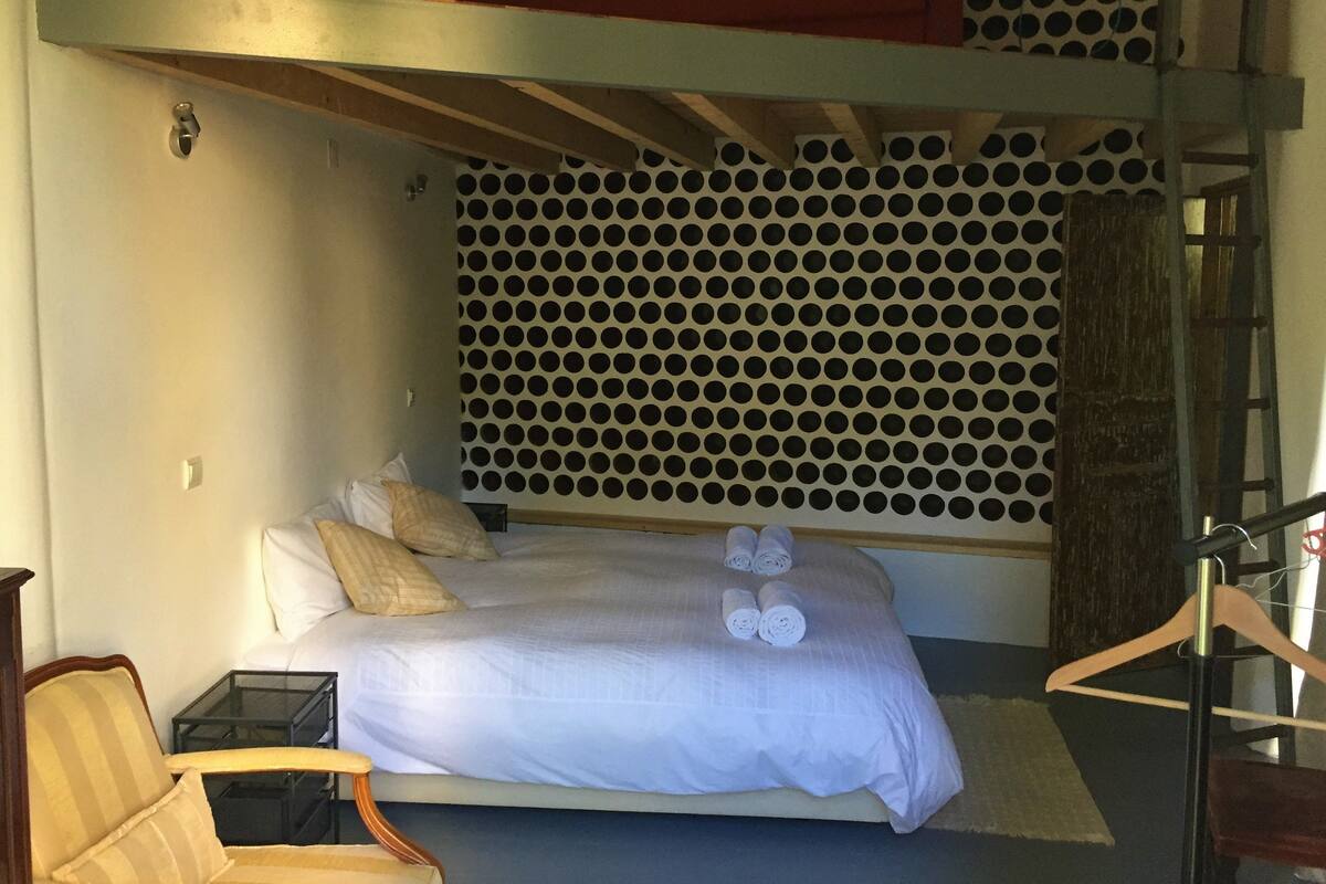 A bedroom with a bunk bed, a bed with rolled towels, a chair, and a patterned wall.