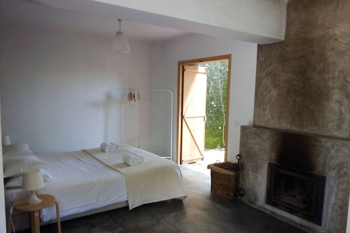 A bedroom with a bed, a bedside table, a lamp, a mirror, a fireplace, and a door leading to an outdoor area.