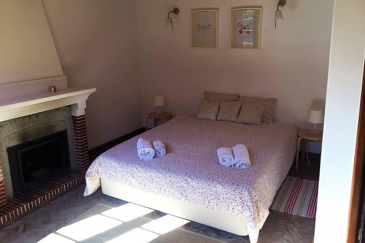 A bedroom with a fireplace, a bed with floral bedding, two wall-mounted lamps, and a framed butterfly picture.