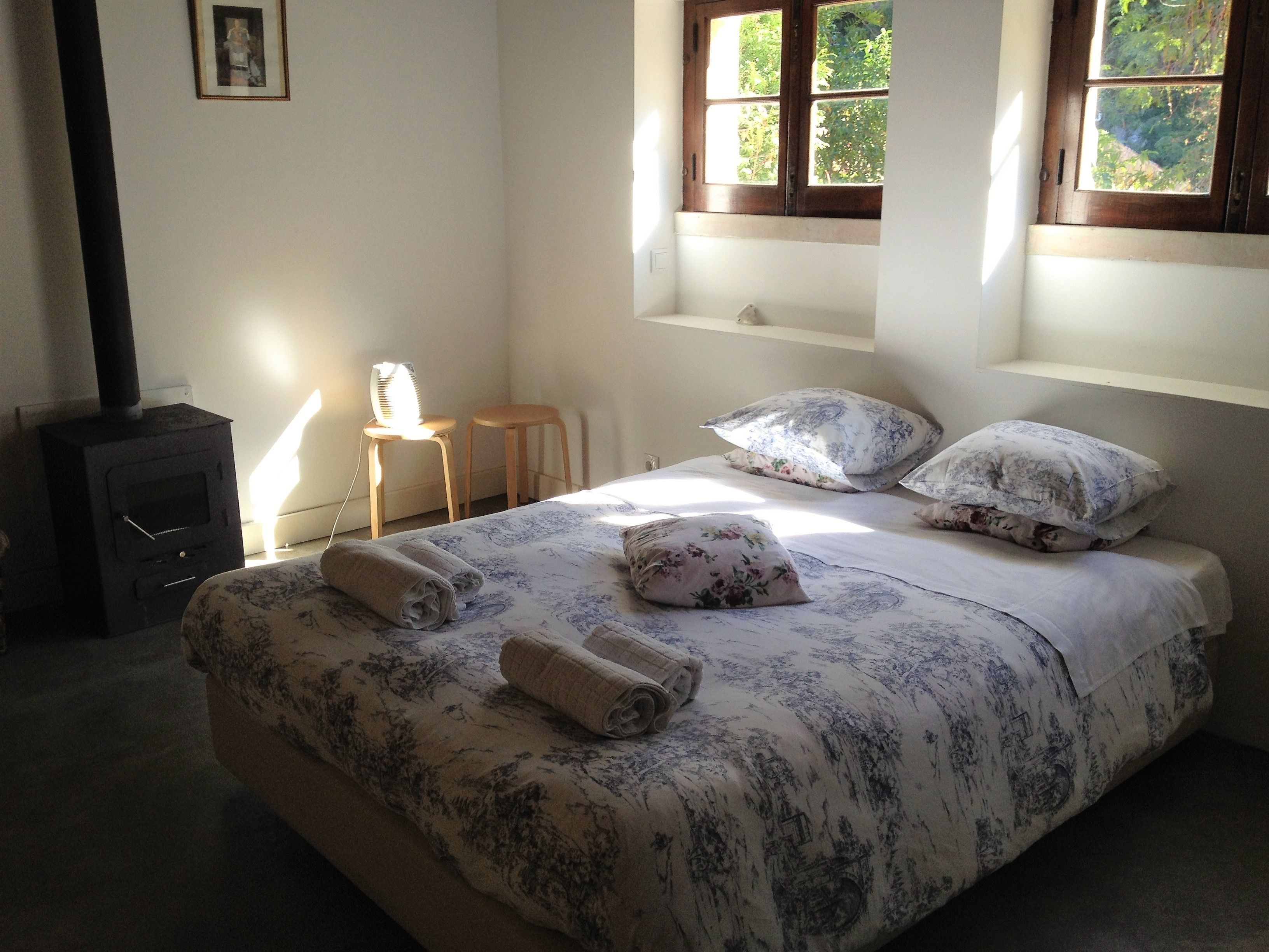 Un dormitorio con cama, dos almohadas, mesita de noche, una silla, calefactor y ventana con vista a follaje.