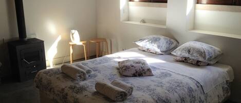 Un dormitorio con cama, dos almohadas, mesita de noche, una silla, calefactor y ventana con vista a follaje.
