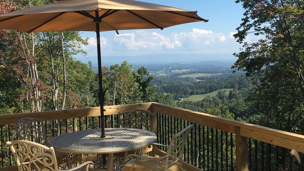 Una terraza de madera con una mesa redonda y sillas, una sombrilla y una vista panorámica de un valle y montañas.