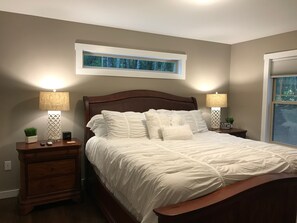 A bedroom with a wooden bed, two bedside tables with lamps, a nightstand, a small potted plant, and a window with a view of trees.