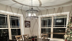 Un comedor con mesa de madera, sillas y un banco. Hay una araña colgando sobre la mesa, y un árbol de Navidad a la derecha.