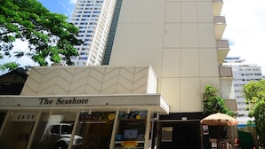 Un edificio alto y moderno con fachada color beige y varios balcones. En primer plano, una tienda con un letrero que dice "The Seashore".
