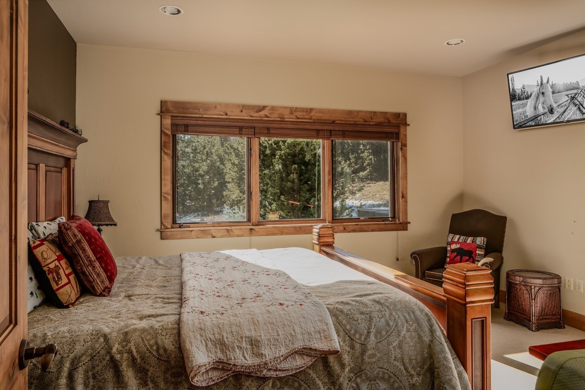 A bedroom with a large window, a bed with a floral bedspread, a chair with a moose pillow, and a small side table.