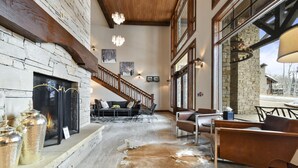 A spacious living area with a stone fireplace, a staircase, and large windows offering a view of the outdoors.