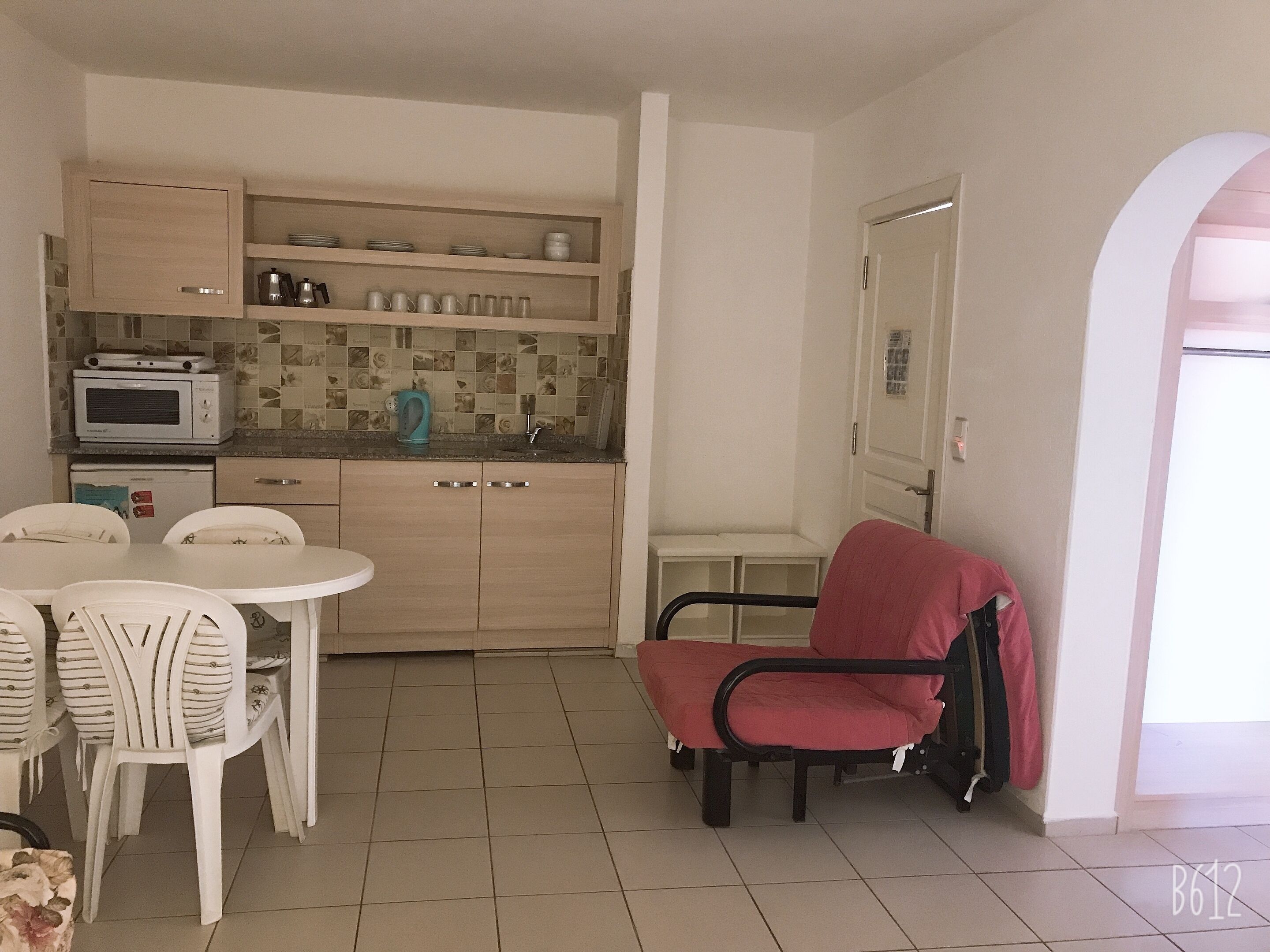A compact kitchen with a microwave, coffee maker, and a tiled backsplash. There is a dining table with chairs and a red chair in the corner.
